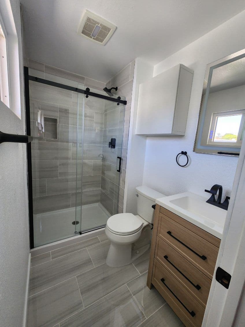 Modern bathroom with glass shower, wooden vanity, and gray tile flooring.